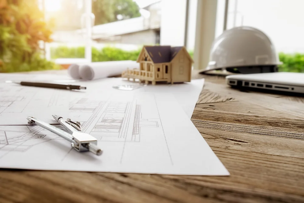Imagem de mesa com planta baixa, com casinha pequena, notebook e capacete EPI
